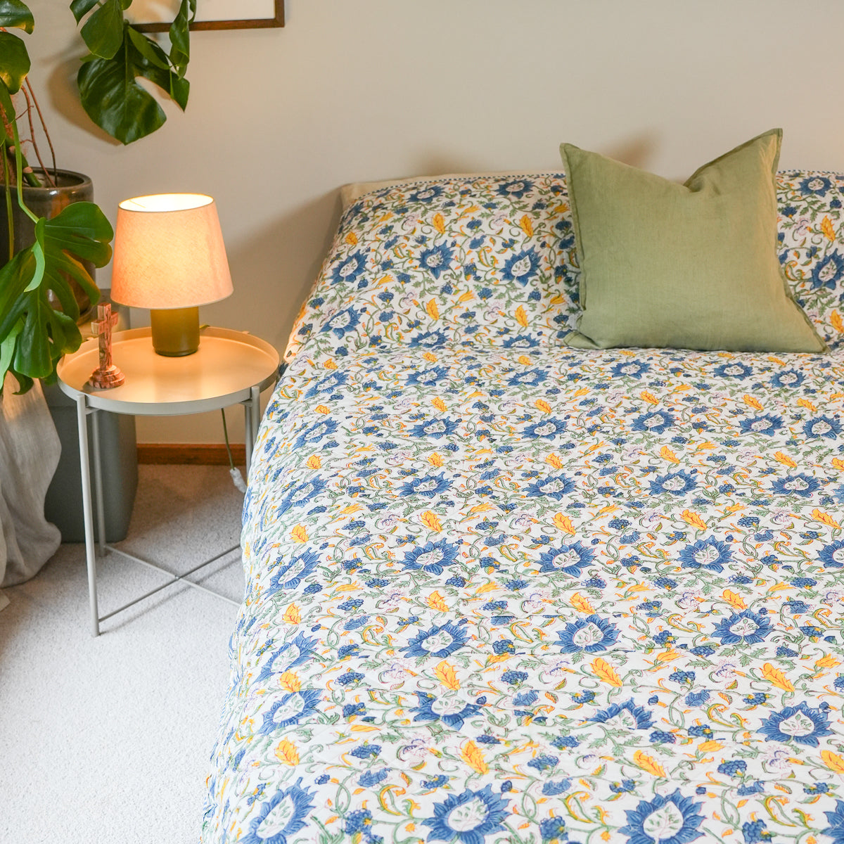 A floral quilted comforter with a blue, yellow, and white pattern styled on a bed in a bedroom setting, featuring a green pillow and a bedside table with a lit lamp.