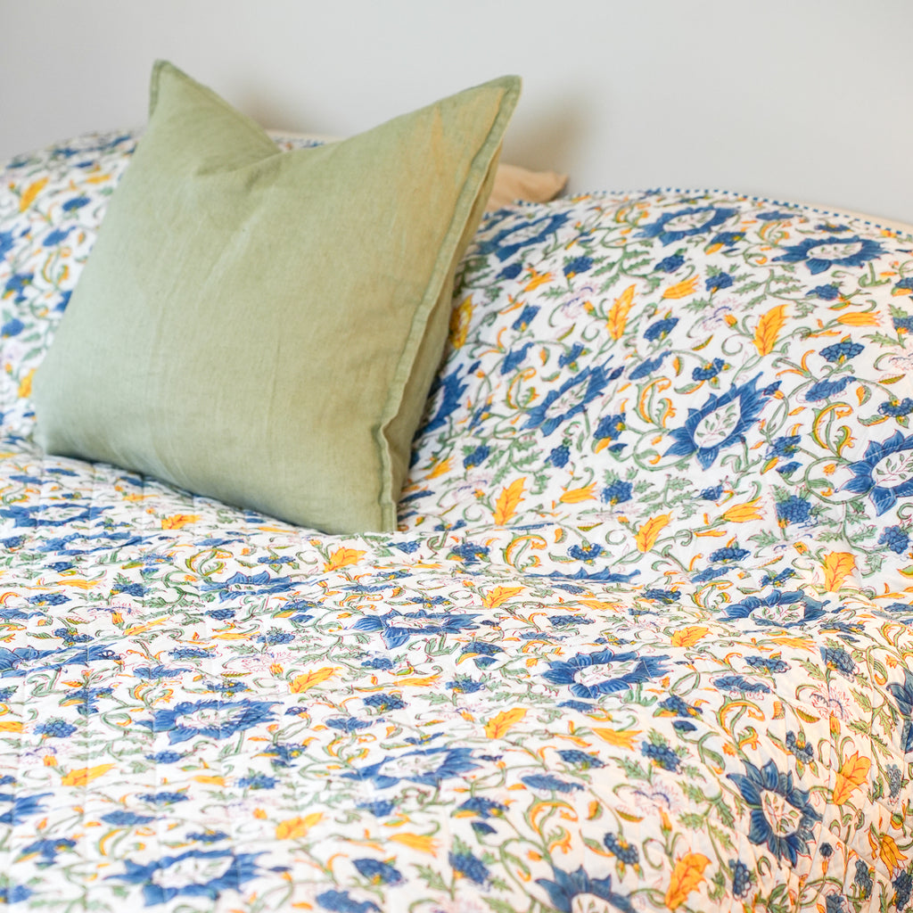 A close-up of a bed or sofa covered with a white quilt or throw featuring an allover floral pattern in blue, yellow, and green, with a solid sage green pillow in the foreground.