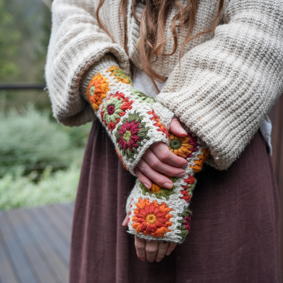 A person wearing a cream knit sweater and brown skirt holds up a pair of chunky crocheted arm warmers decorated with colorful floral motifs in orange, pink, and green.
