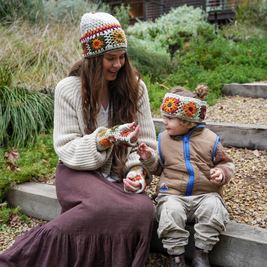 A woman and a child sitting outdoors on stone steps, both wearing matching cream knit beanie and mitten sets with orange, yellow, and red floral patterns. The woman wears a cream knit sweater and a dark brown skirt, while the child wears a tan puffer vest, beige pants, and dark boots.