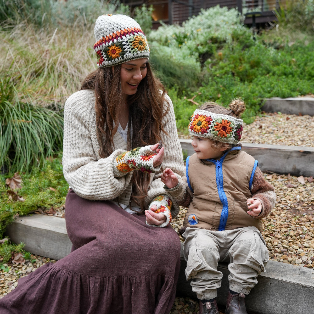 A woman and a child sitting outdoors on stone steps, both wearing matching cream knit beanie and mitten sets with orange, yellow, and red floral patterns. The woman wears a cream knit sweater and a dark brown skirt, while the child wears a tan puffer vest, beige pants, and dark boots.