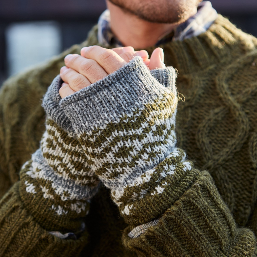 Close-up of a person wearing olive green and white patterned knit fingerless gloves over an olive green cable-knit sweater, with hands clasped together.