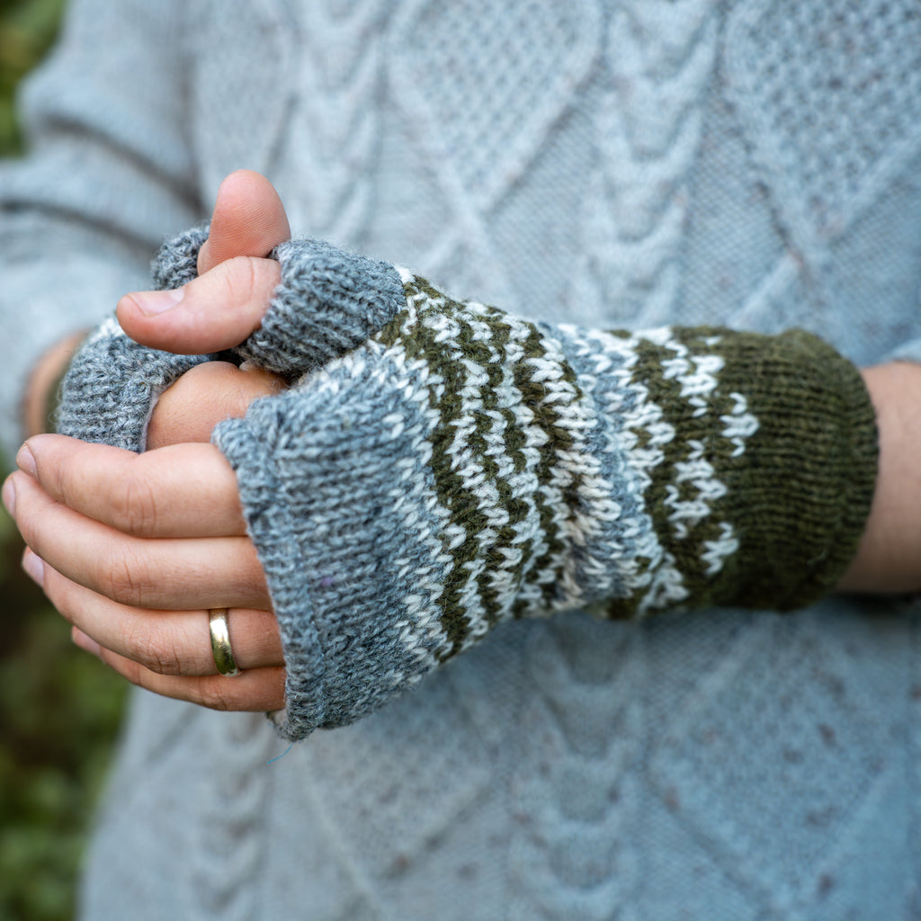 Close-up of a person wearing a pair of fingerless knit wrist warmers with a blue-gray base, dark green and white Fair Isle pattern, and olive green cuffs, shown against a light blue cable-knit sweater.