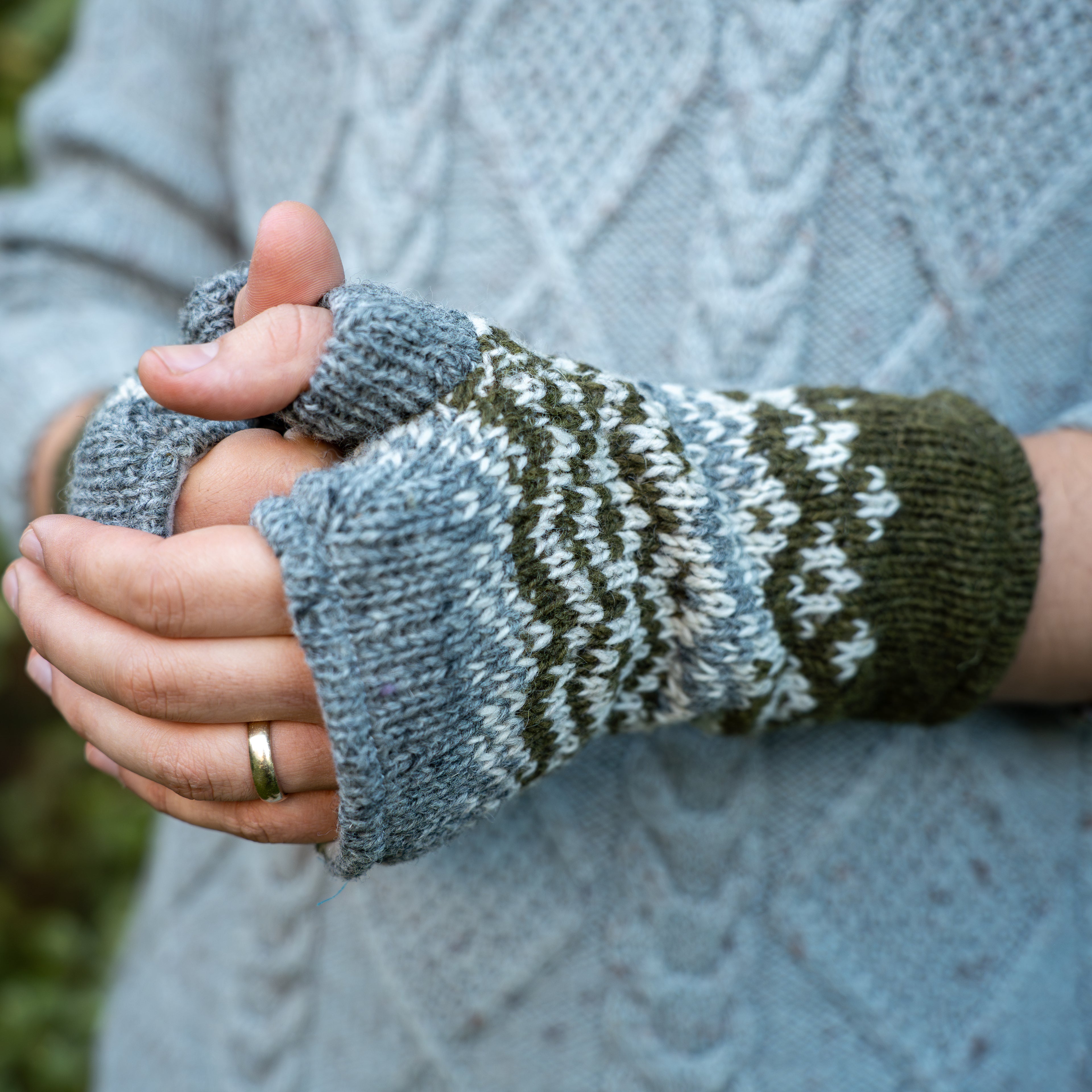 Close-up of a person wearing a pair of fingerless knit wrist warmers with a blue-gray base, dark green and white Fair Isle pattern, and olive green cuffs, shown against a light blue cable-knit sweater.