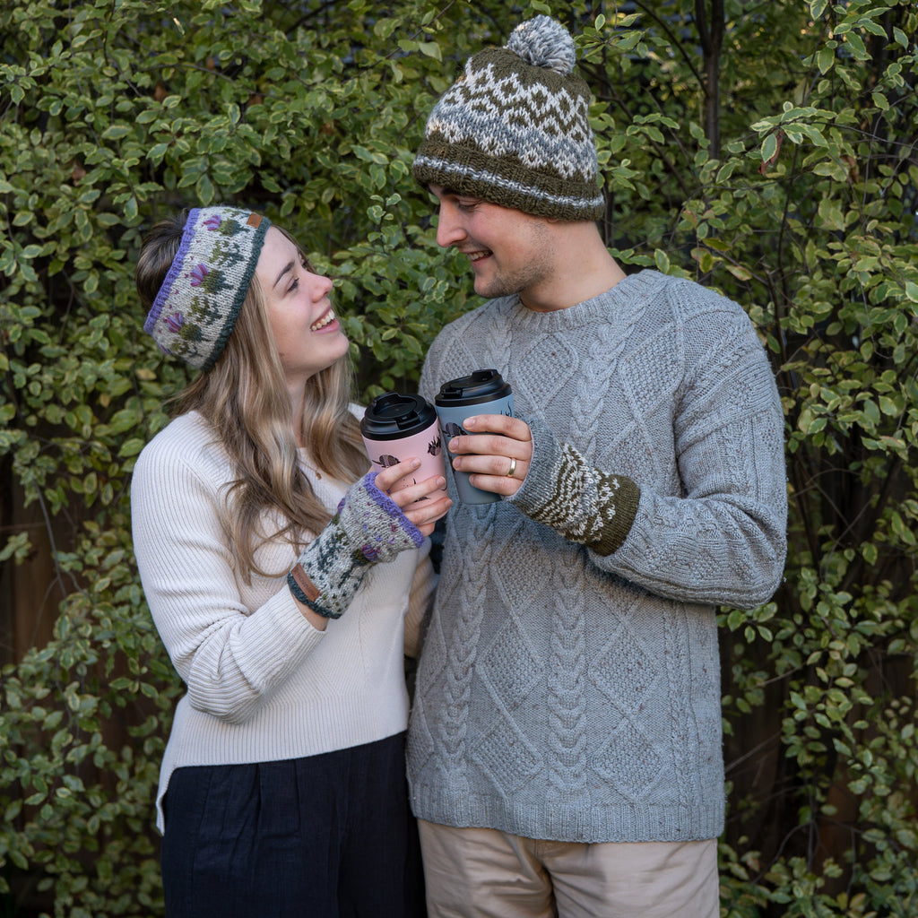 A couple outdoors in front of green foliage, wearing matching knit winter accessories including a patterned beanie, matching knit gloves, and a cream knit sweater. The man wears a gray cable-knit sweater and beige pants, while the woman wears a cream knit sweater and dark pants.