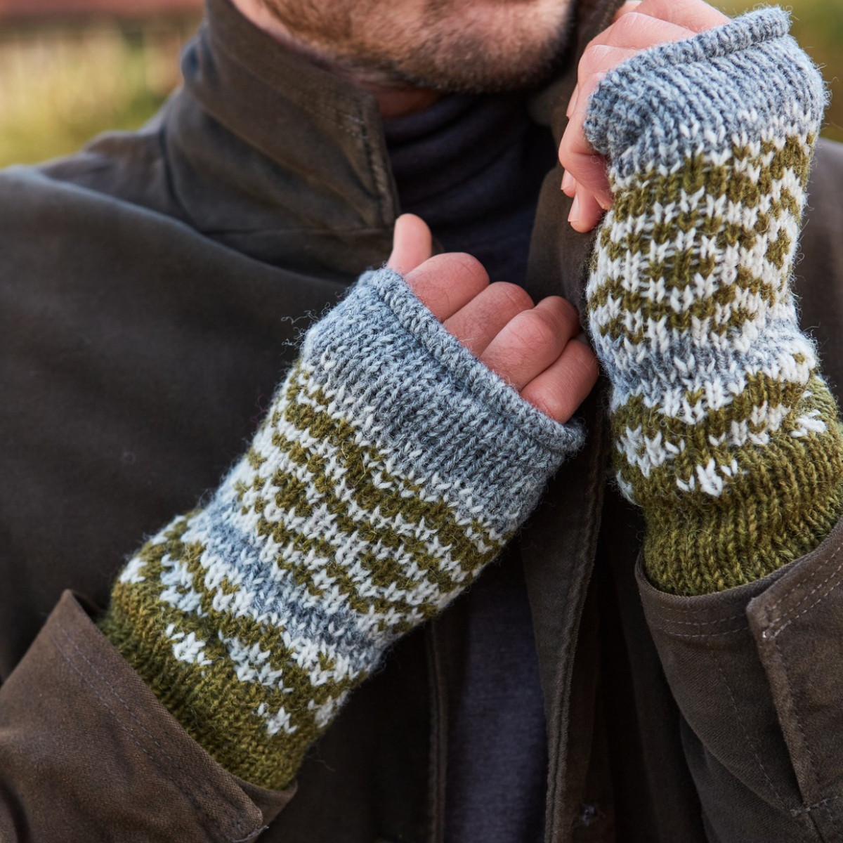 Close-up of a person wearing hand-knit fingerless gloves with a blue-gray and olive green patterned design, paired with a brown jacket.