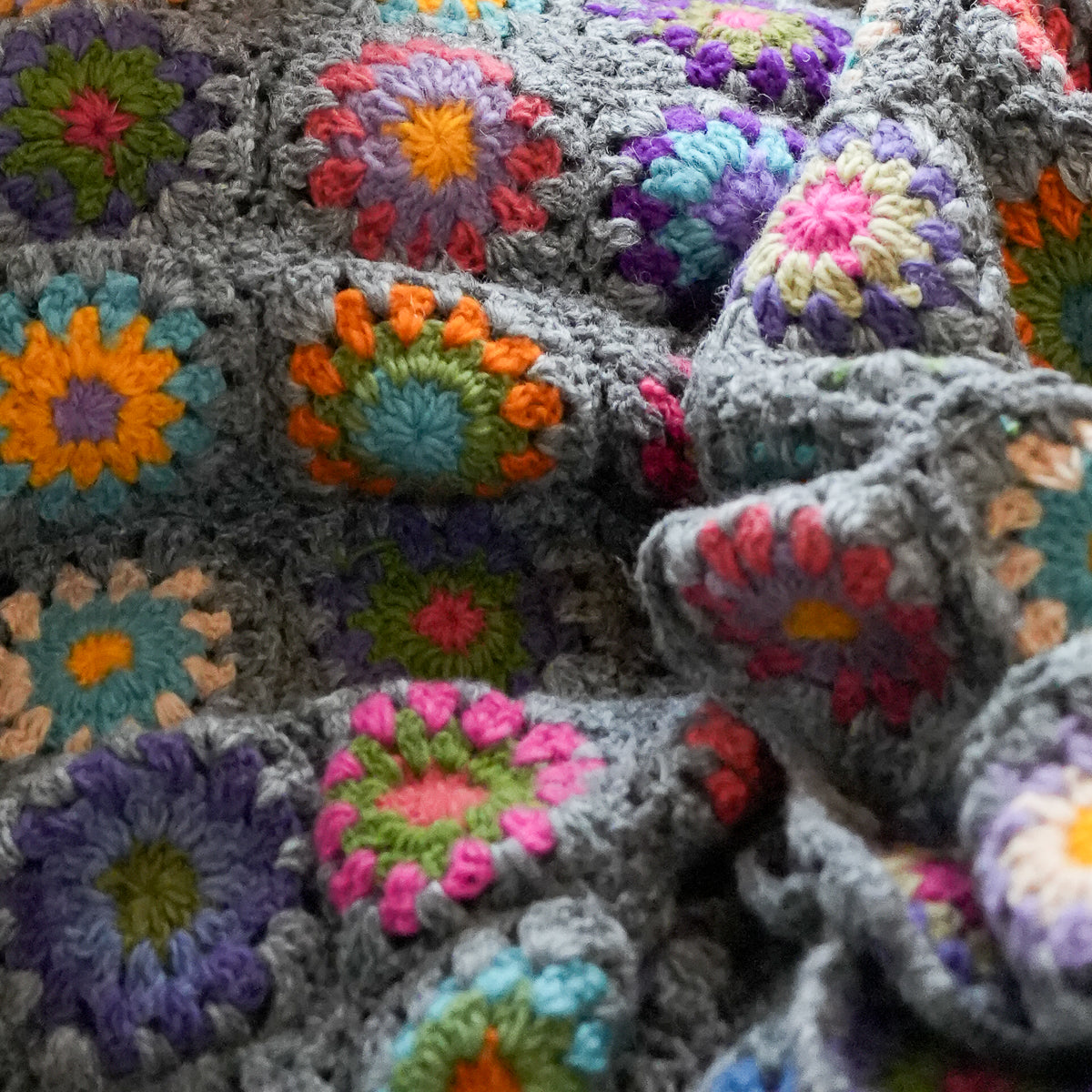 A close-up of a handmade crocheted blanket featuring a gray base with colorful granny square flowers in pink, orange, purple, blue, green, and yellow.