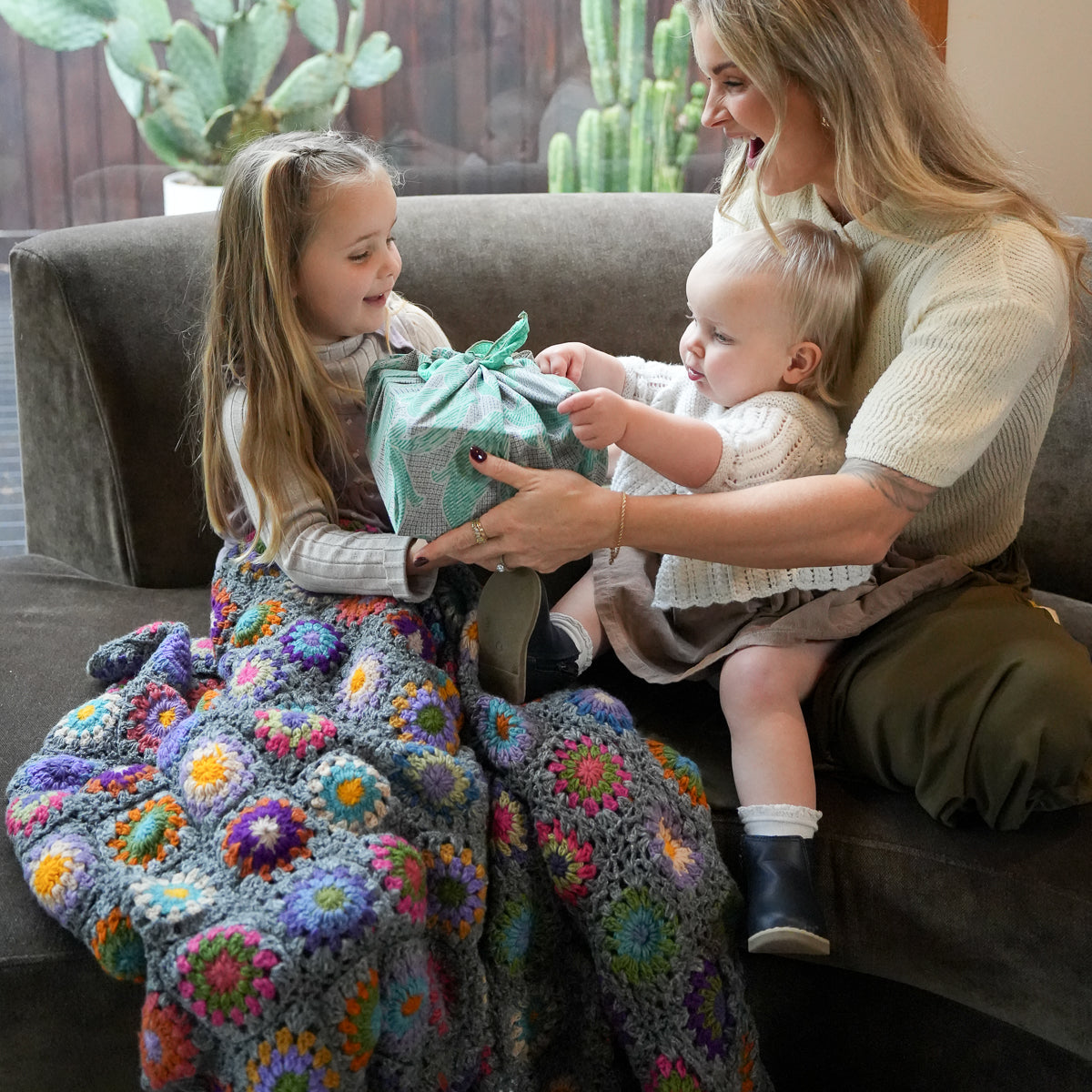 A woman and a young girl sit on a gray couch, smiling as the woman holds a baby while wrapping a gift box. The girl is draped in a colorful crocheted blanket with large floral patterns in purple, blue, yellow, green, and pink.
