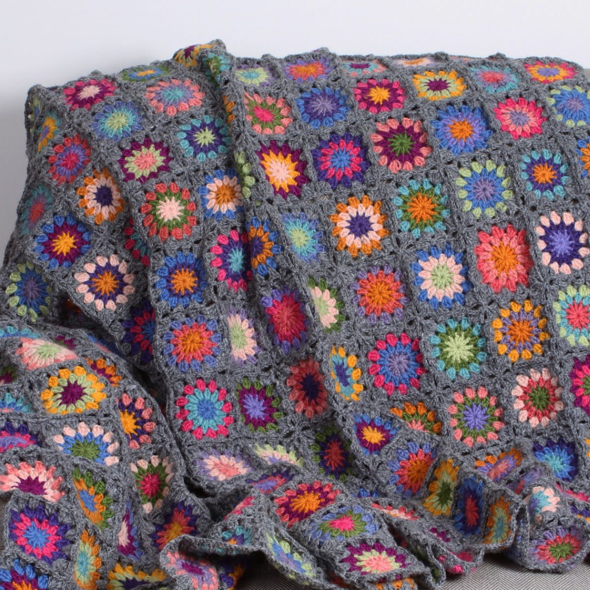 A handmade crocheted throw blanket featuring a gray yarn base with colorful granny square floral motifs, draped over a surface against a plain light gray background.