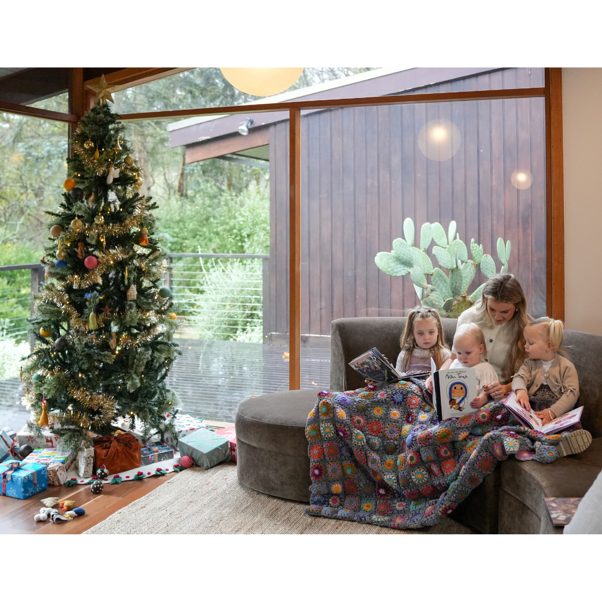 A multicolor patchwork quilted blanket is draped over a gray sofa in a cozy living room, with a woman and two young children reading to them. A decorated Christmas tree and wrapped gifts are visible in the background near large windows.