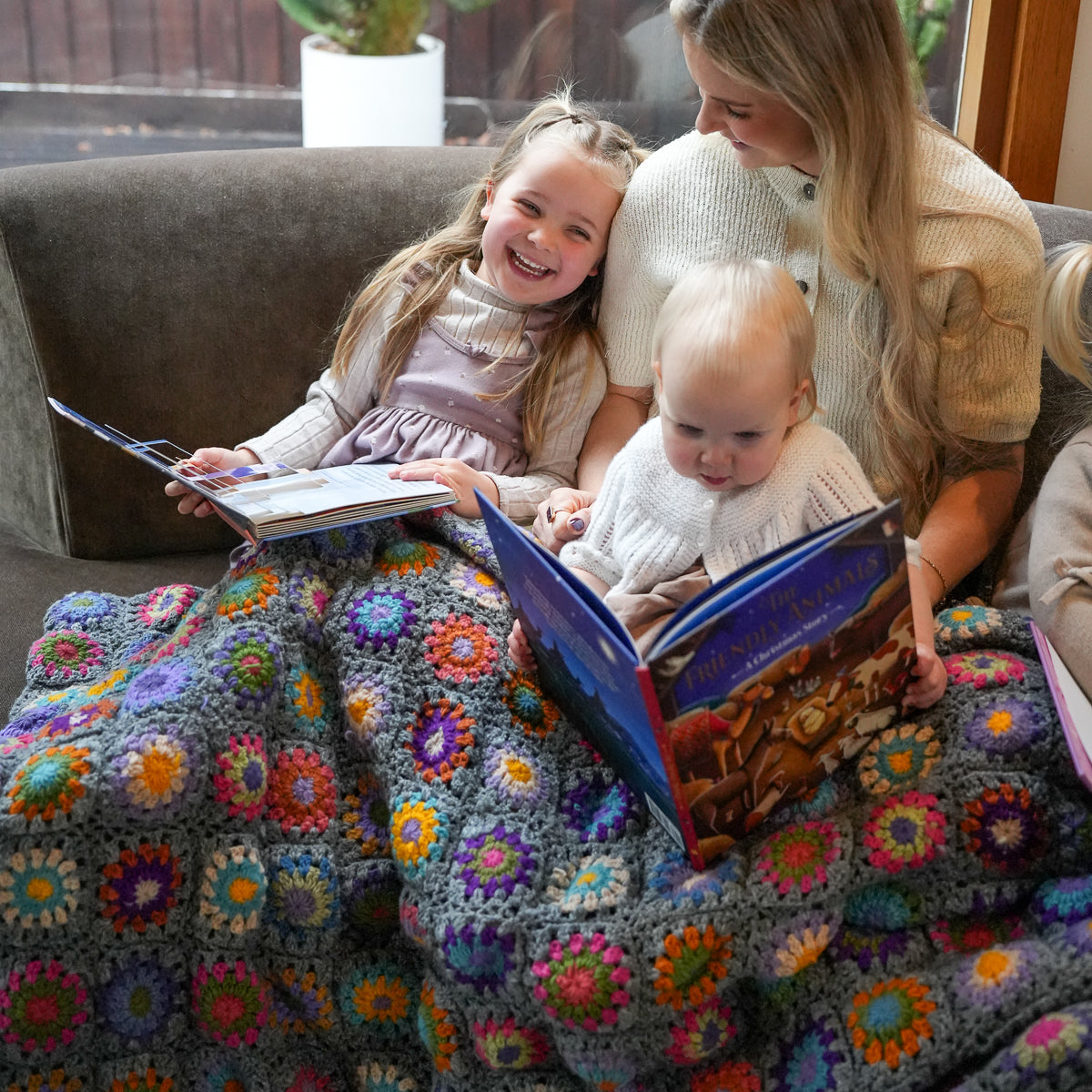 A woman and two children sit on a couch reading books under a colorful crocheted blanket with a gray base and multicolored floral granny-square patterns.