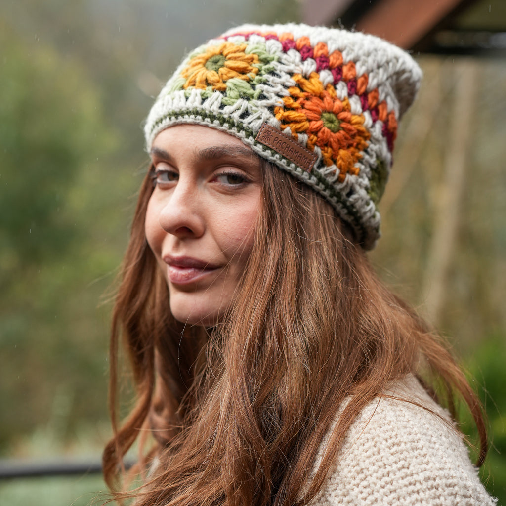 A woman wearing a handmade crocheted slouchy beanie with colorful floral patterns in orange, green, yellow, and purple on a cream base, set against a blurred outdoor background.