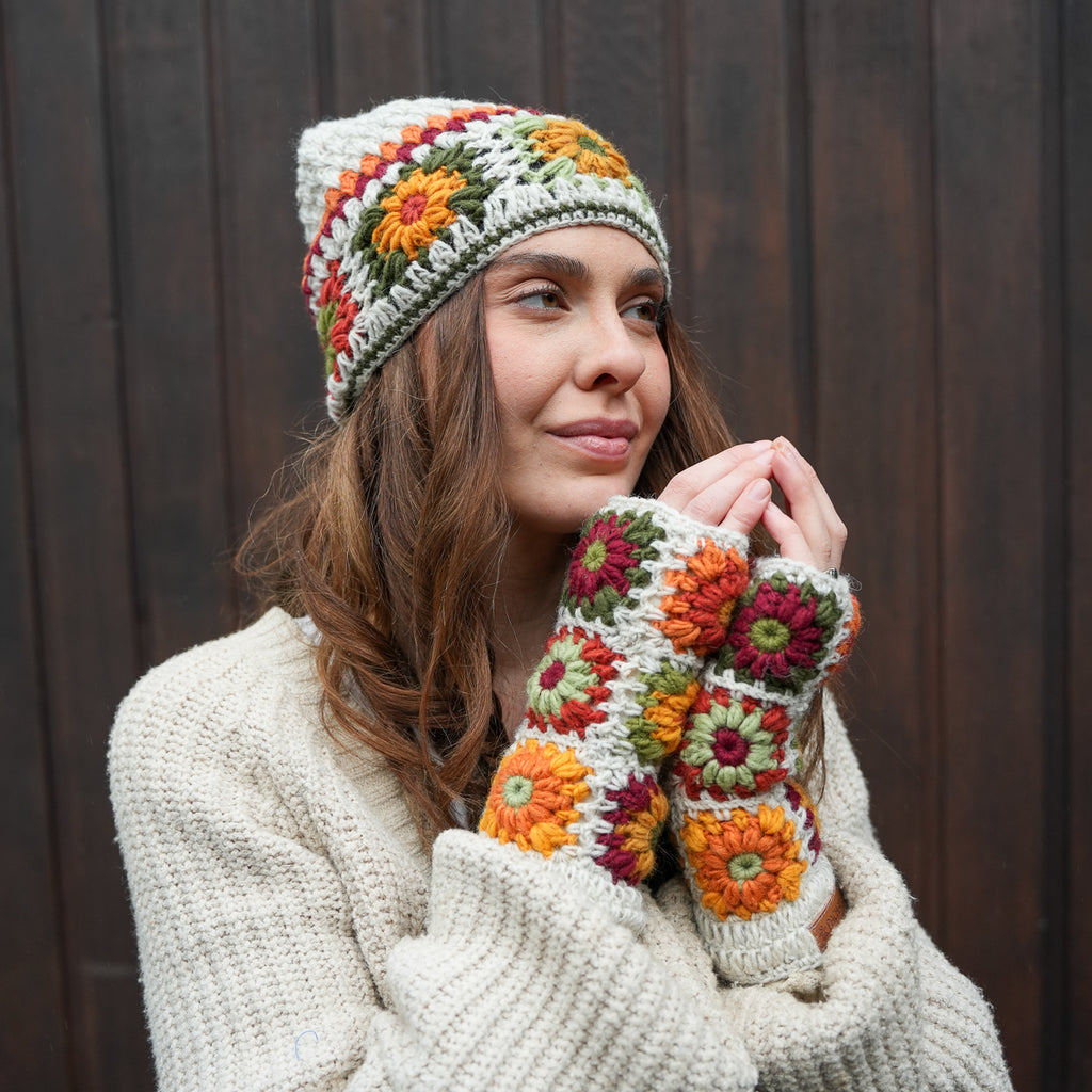 A woman wearing a cream-colored crochet beanie and matching fingerless gloves with colorful granny square floral patterns in orange, green, and red, posed against a dark wooden background.