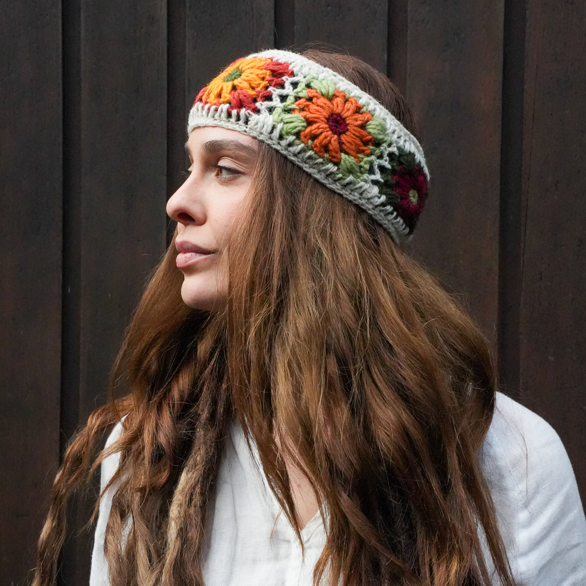 A woman with long wavy brown hair wearing a handmade crocheted headband with colorful floral patterns, posed against a dark wooden background.