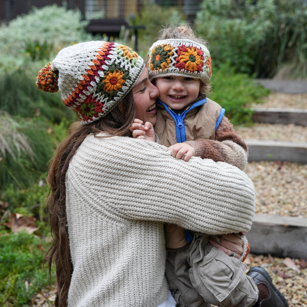 A woman and a young child outdoors, both wearing handmade crocheted knit beanies with colorful floral and geometric patterns. The woman wears a cream ribbed sweater, and the child wears a tan jacket with a blue zipper.