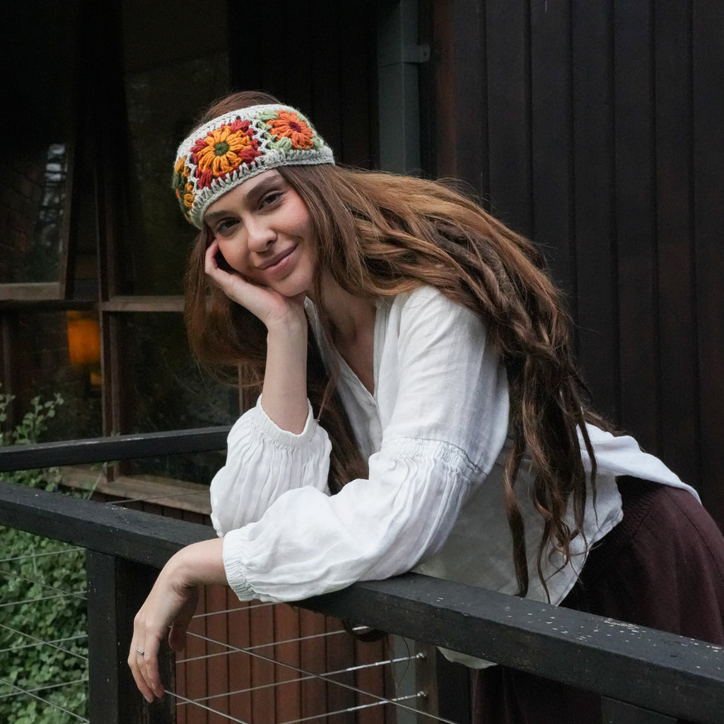 A woman with long brown hair leans on a dark railing in front of a wooden building, wearing a white blouse and a colorful crocheted headband with orange, yellow, and red floral patterns.
