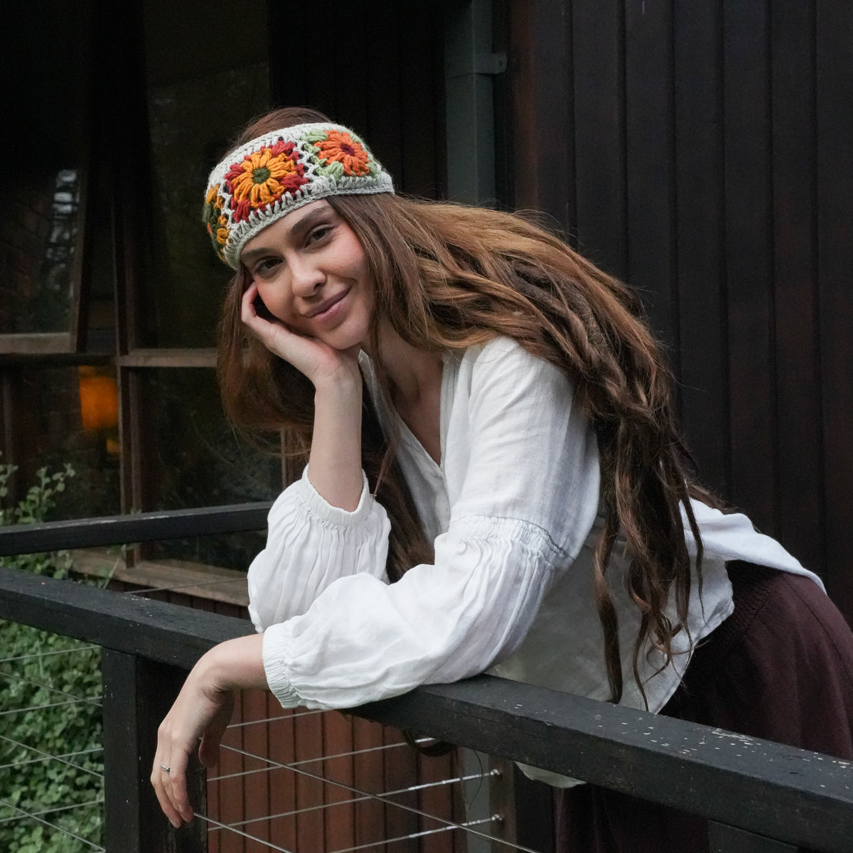A woman with long brown hair leans on a dark railing in front of a wooden building, wearing a white blouse and a colorful crocheted headband with orange, yellow, and red floral patterns.
