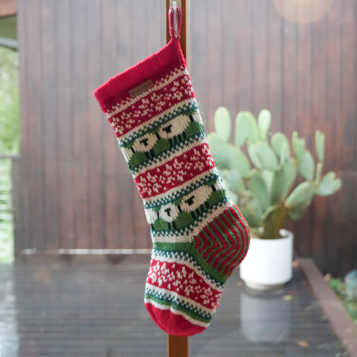 A red, green, and cream knit Christmas stocking with a snowman pattern hangs from a wooden pole on a wet wooden deck, with a potted cactus in the background.