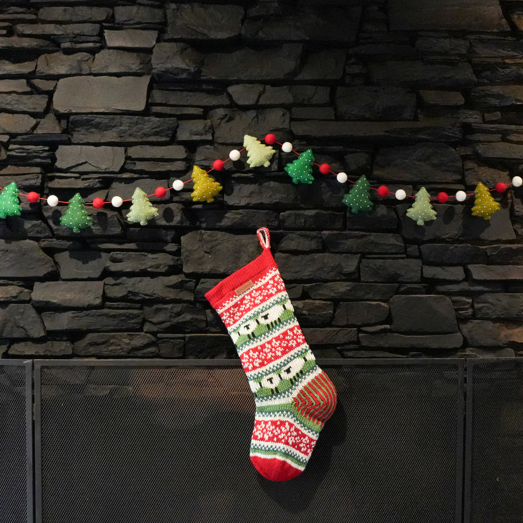 A festive Christmas stocking with a red, green, and white fair isle pattern hangs on a black fireplace screen in front of a dark gray stone wall. A decorative string of red, white, green, and yellow pom-pom Christmas trees and beads is strung across the wall above.