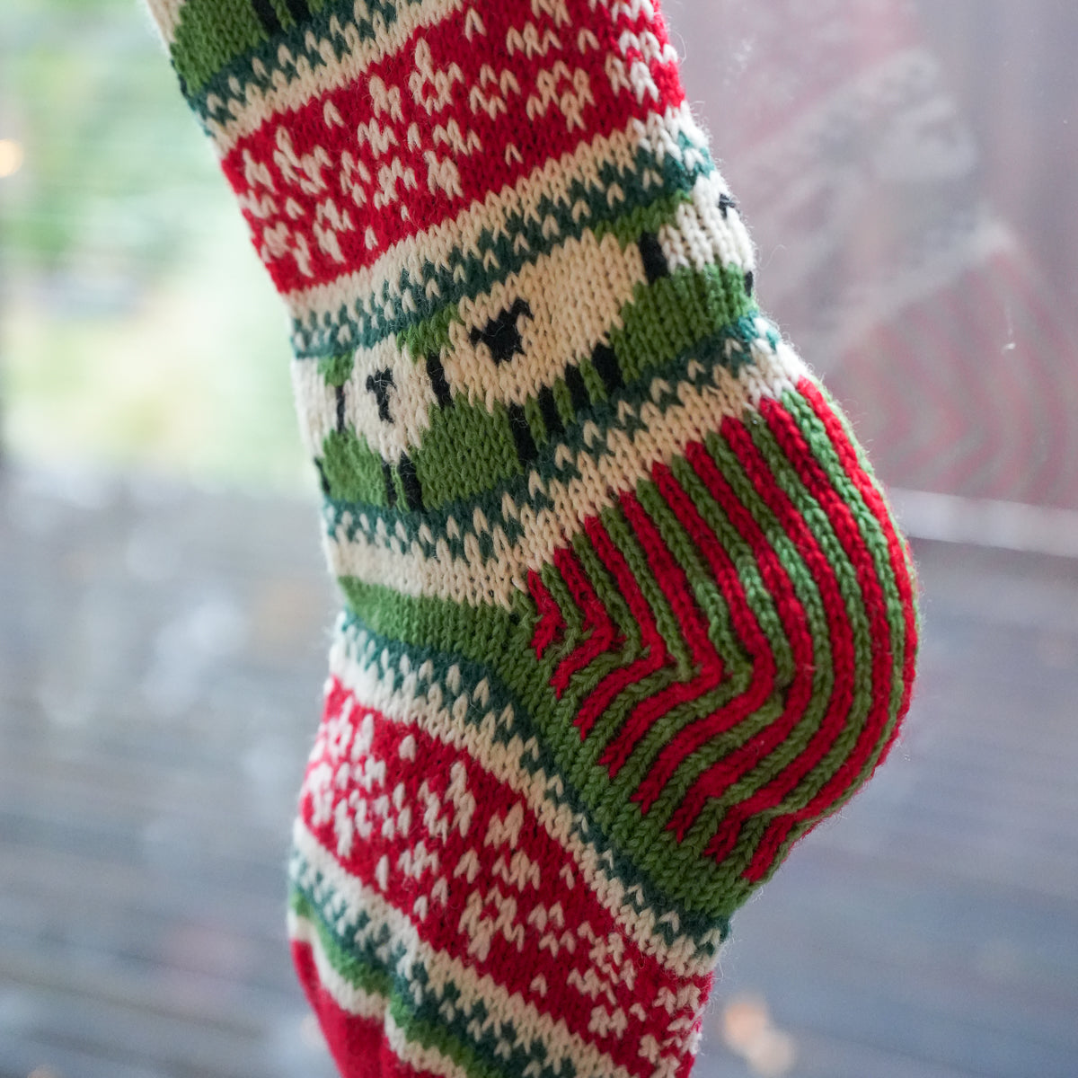 A close-up of a hand-knit Christmas stocking with festive patterns in red, green, and cream, hanging near a window.