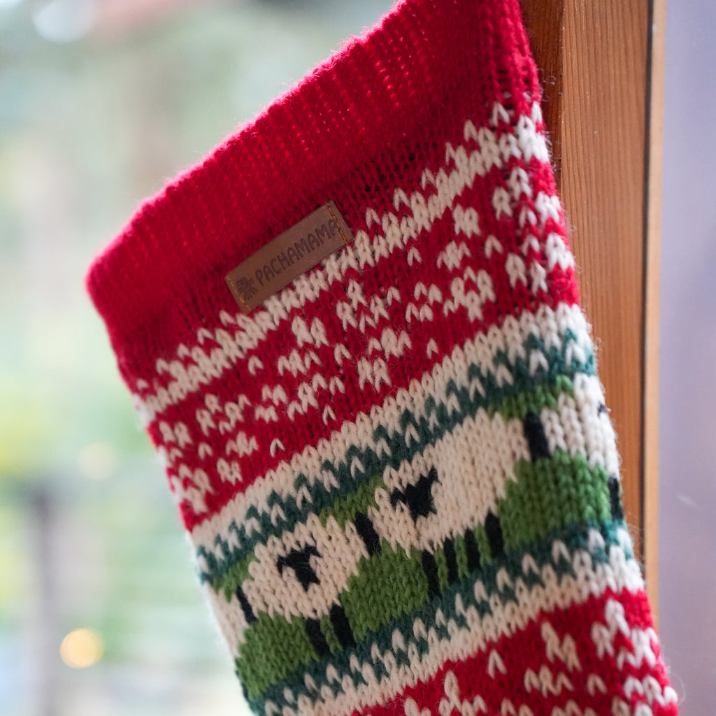 A close-up of a knitted Christmas stocking with a red cuff, cream and red patterned stripes, and green and cream animal motifs, hanging near a wooden frame with a blurred outdoor background.