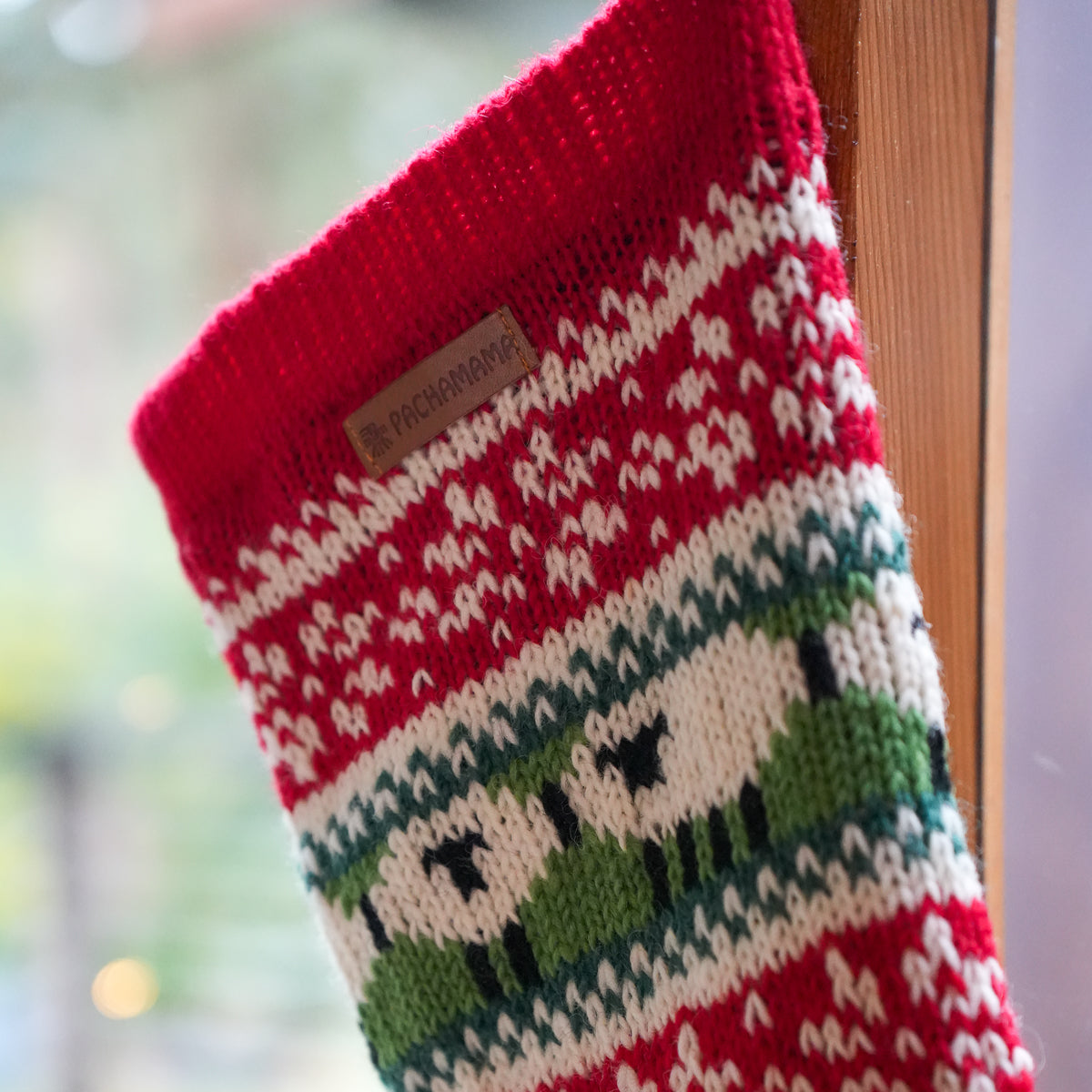 A close-up of a knitted Christmas stocking with a red cuff, cream and red patterned stripes, and green and cream animal motifs, hanging near a wooden frame with a blurred outdoor background.