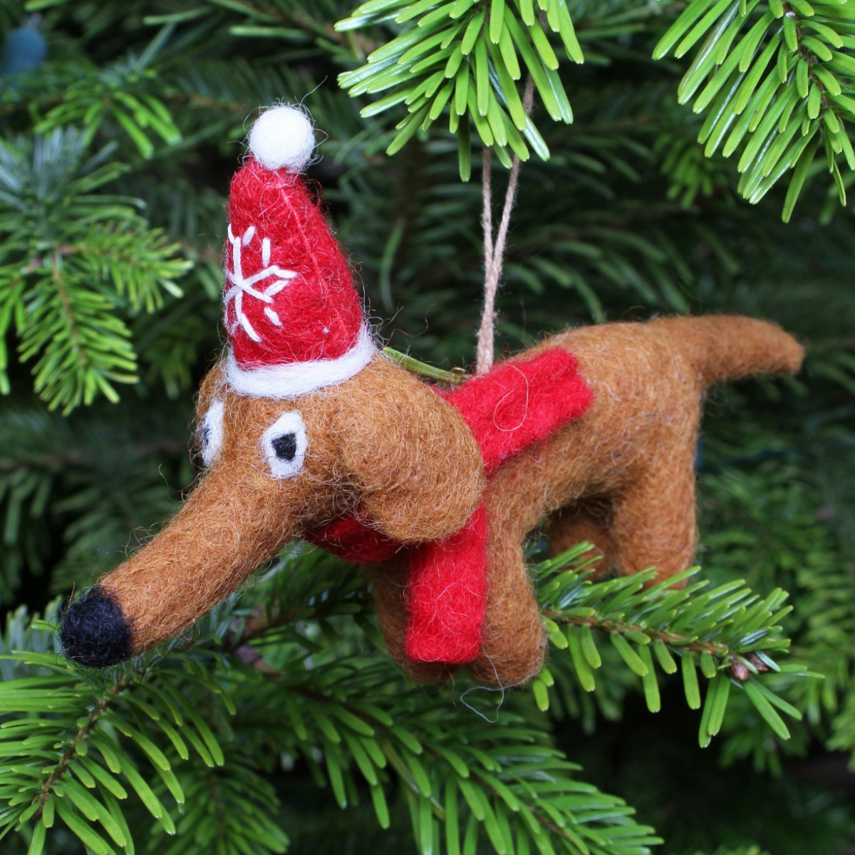 A brown felt dog Christmas ornament wearing a red Santa hat and red scarf, hanging from a string in a green pine tree.