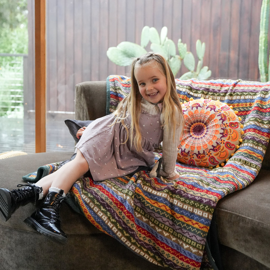 A young girl with long blonde hair sits smiling on a brown couch, wearing a mauve dress and black boots. The couch is styled with a vibrant, multicolored patterned throw blanket and a matching round mandala pillow in front of a wooden wall background.
