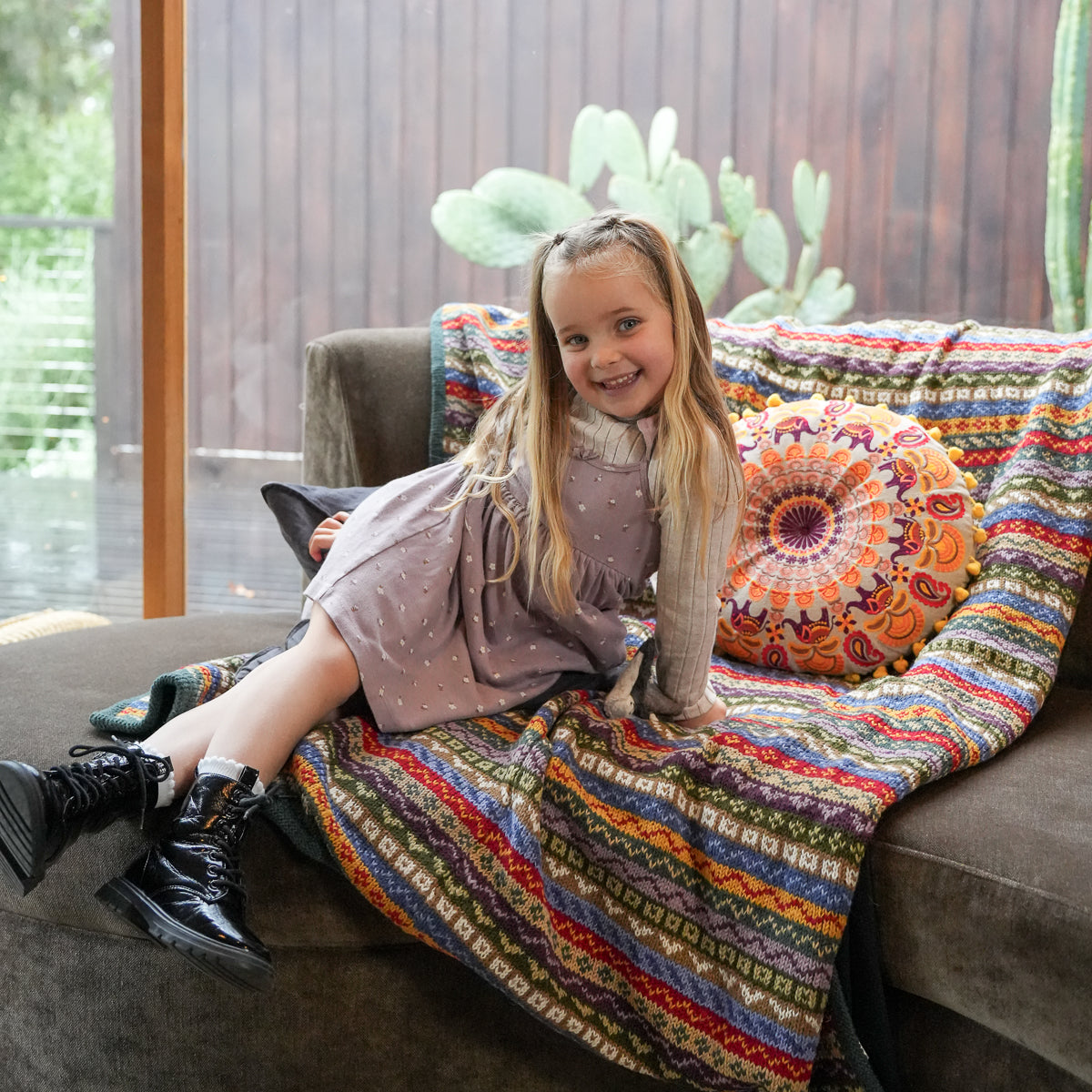A young girl with long blonde hair sits smiling on a brown couch, wearing a mauve dress and black boots. The couch is styled with a vibrant, multicolored patterned throw blanket and a matching round mandala pillow in front of a wooden wall background.