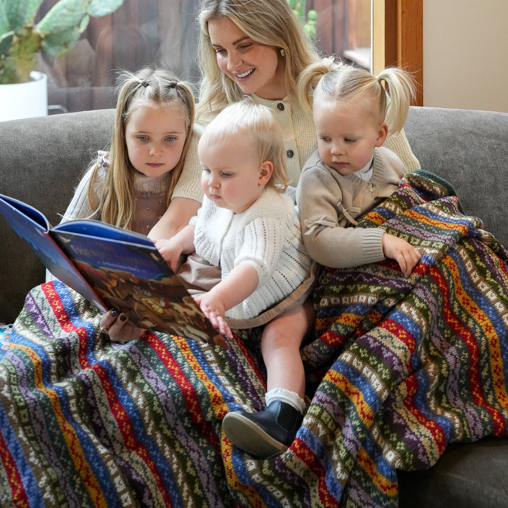 A woman and two young girls sit on a couch reading a book, wrapped together in a thick, multicolored knitted blanket with vertical stripes of red, blue, yellow, green, and purple.