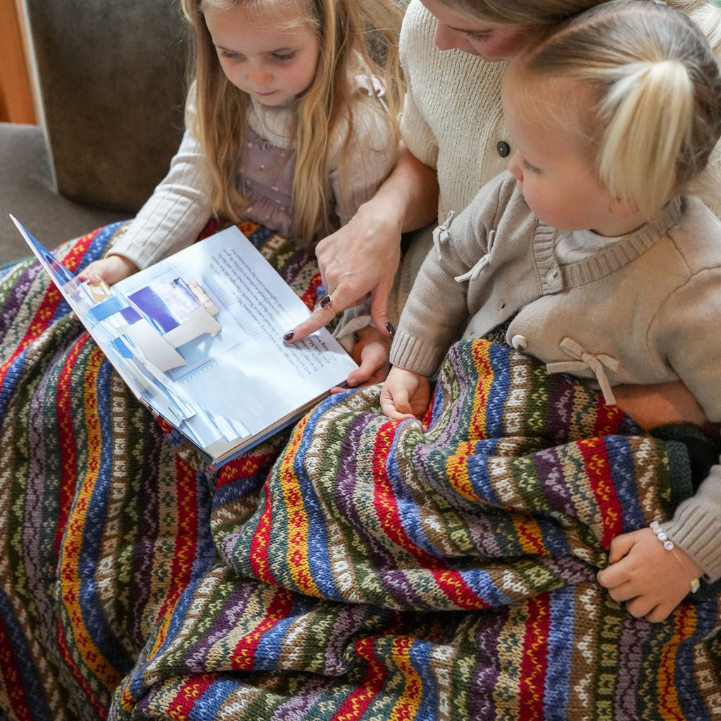 A close-up lifestyle shot of a mother and her two daughters sitting together under a vibrant, multicolored knit blanket while reading a book.