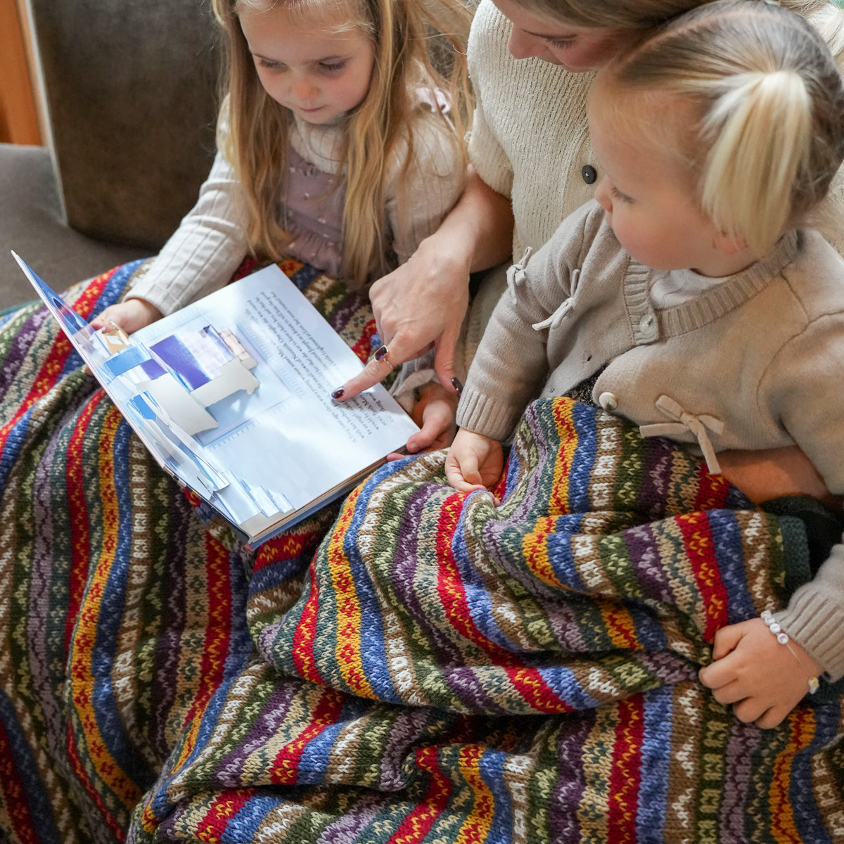 A close-up lifestyle shot of a mother and her two daughters sitting together under a vibrant, multicolored knit blanket while reading a book.