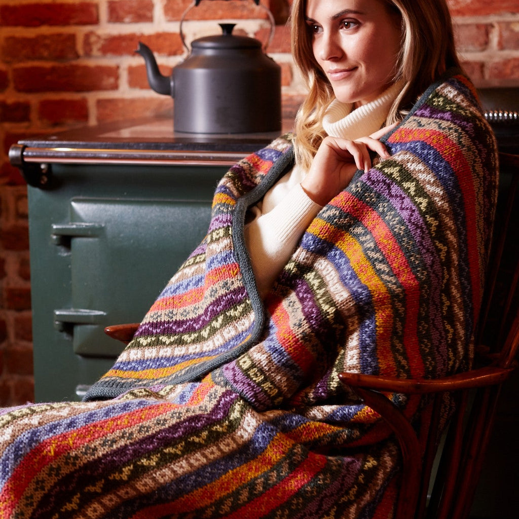 A woman sits in a wooden chair wrapped in a thick, multicolored striped knit blanket, with a dark green stove and a black kettle in the background against a brick wall.