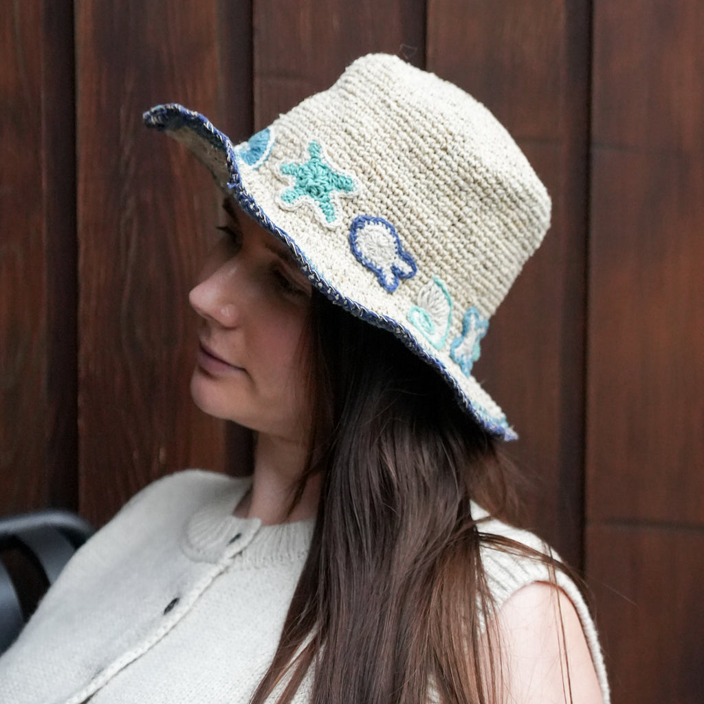 A woman wearing a hand-knitted beige bucket hat with embroidered blue and turquoise sea star and shell motifs, posed against a dark wooden background.
