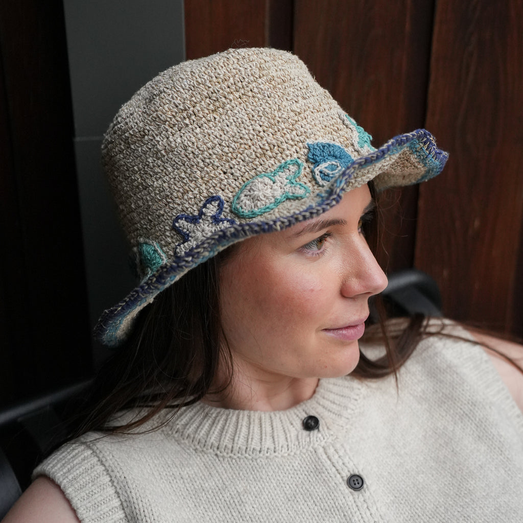 A woman in profile wearing a beige knit bucket hat with blue and green embroidered details and a knitted brim, paired with a matching beige knit top.