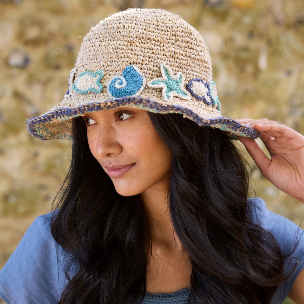 A woman wearing a straw sun hat decorated with blue and green seashell and starfish appliqués, posed outdoors against a blurred natural background.