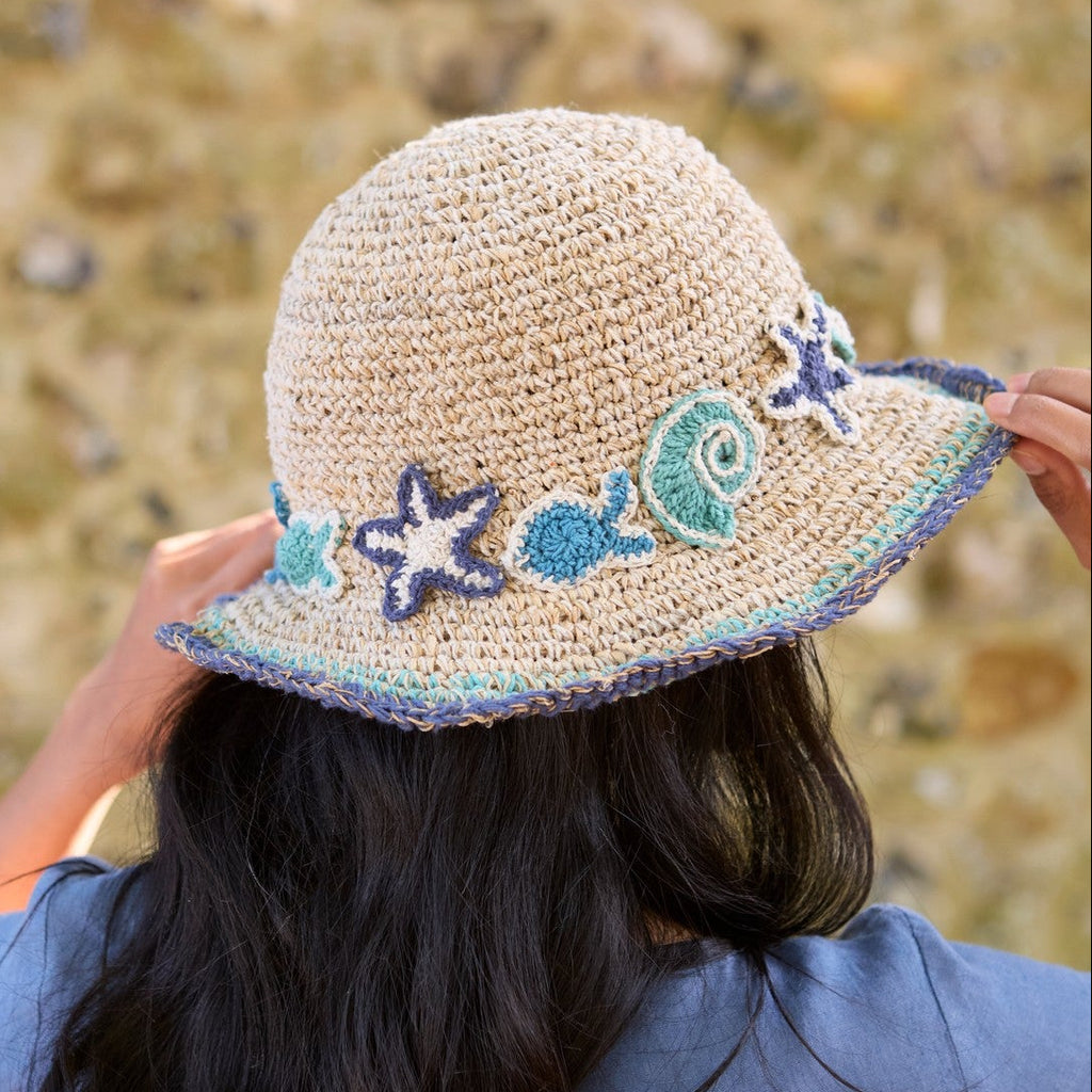A child viewed from behind wearing a beige crocheted sun hat with blue and green sea-themed appliqués, standing outdoors against a blurred stone wall.