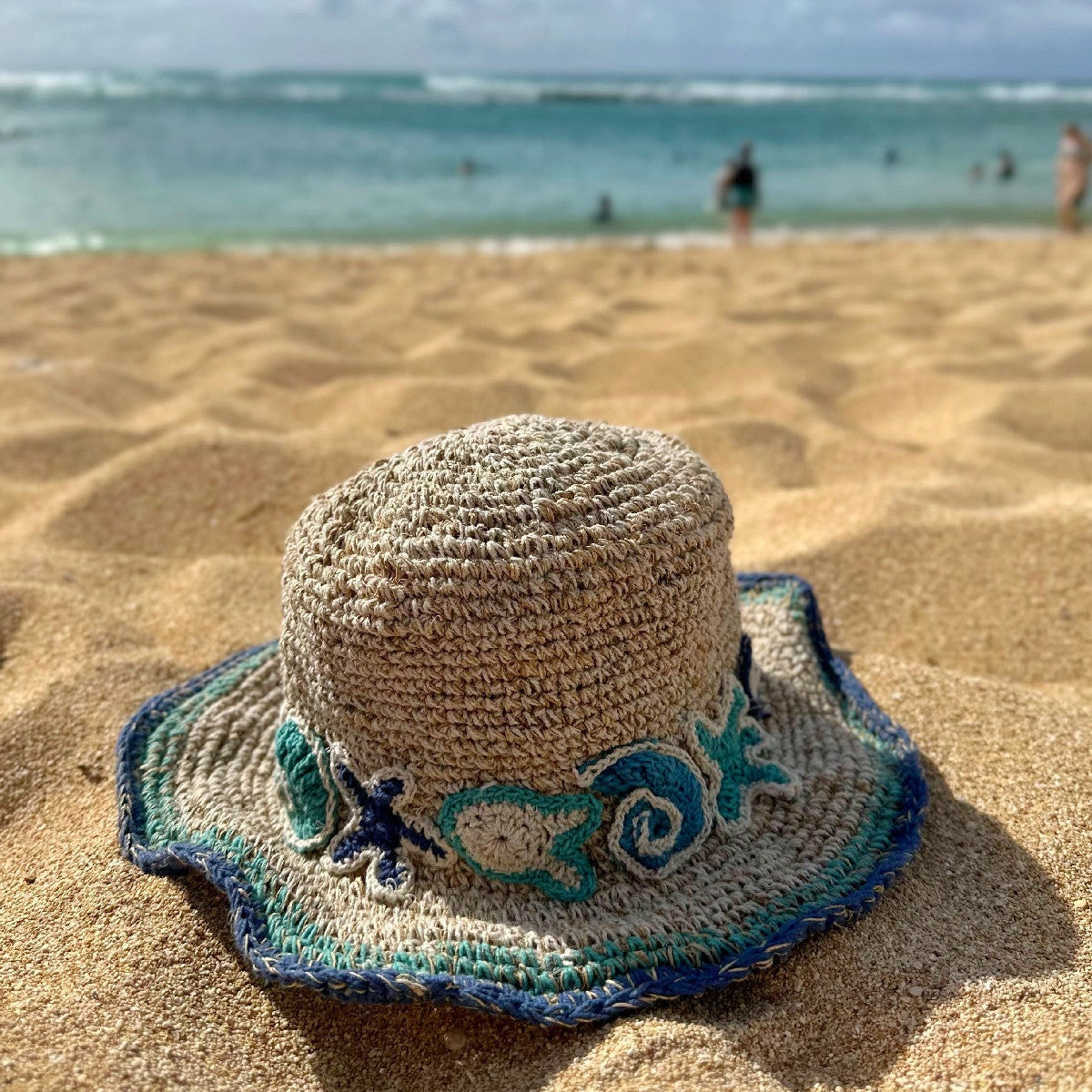 A woven straw sun hat with a blue and green embroidered band, resting on a sandy beach with the ocean and sky blurred in the background.