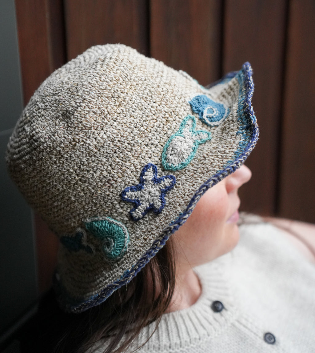 A close-up side profile of a person wearing a chunky knit bucket hat in a heathered beige color, decorated with blue and teal starfish and shell motifs on the brim.