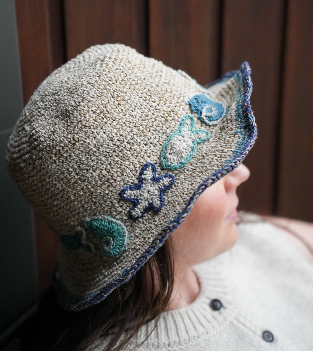 A close-up side profile of a person wearing a chunky knit bucket hat in a heathered beige color, decorated with blue and teal starfish and shell motifs on the brim.