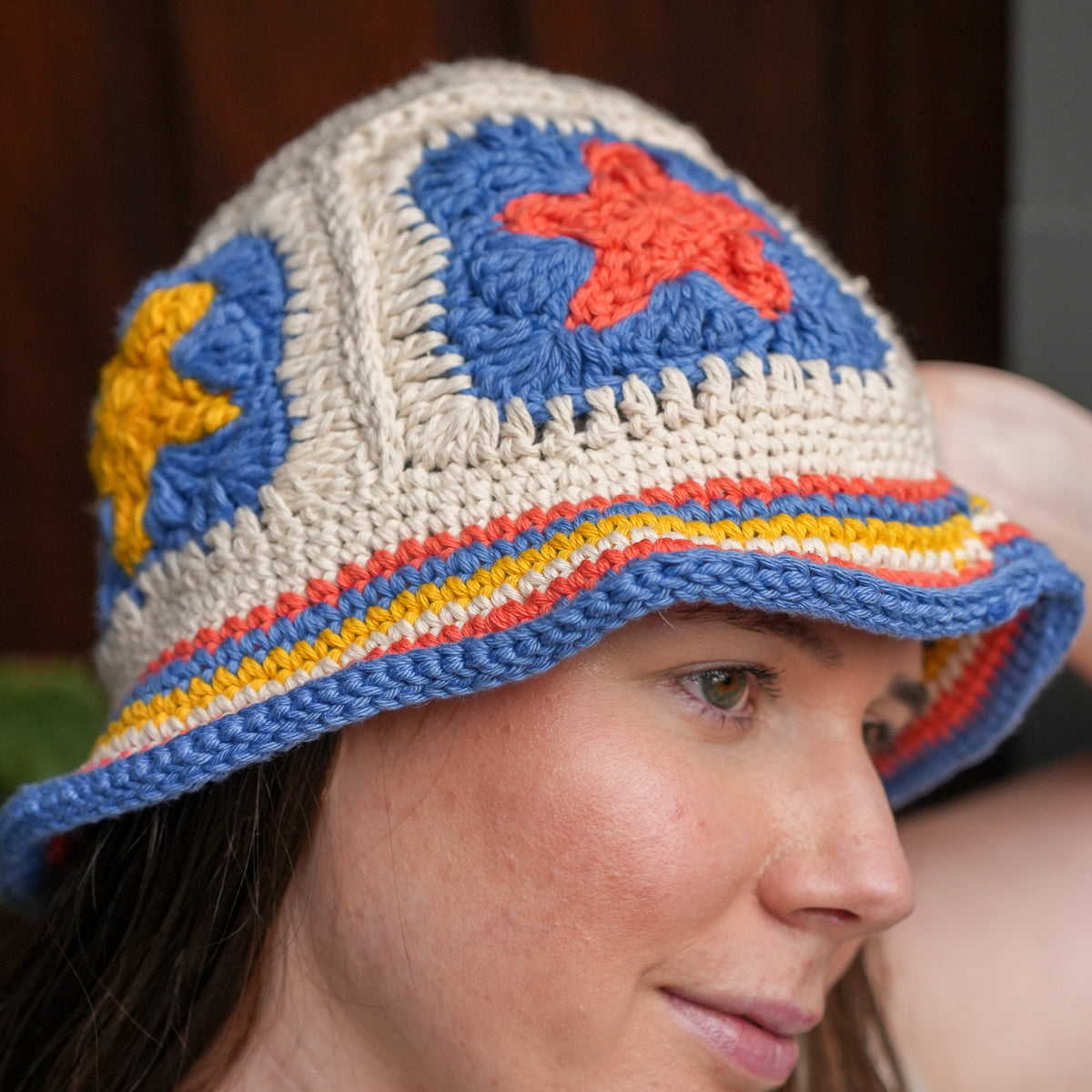 A close-up of a woman wearing a crocheted bucket hat with a cream base and colorful star and stripe details in blue, red, and yellow.