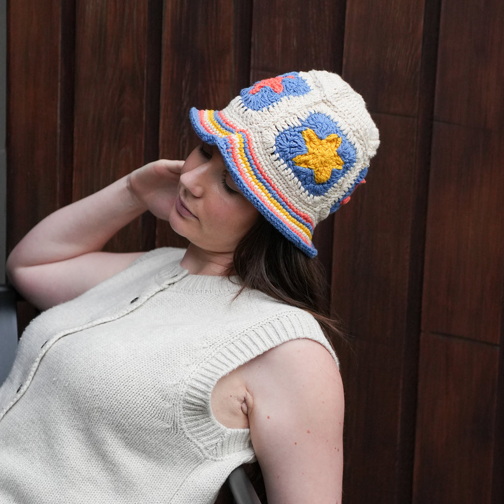 A woman wearing a cream-colored knit bucket hat with colorful crocheted details, including a yellow star, blue and red patches, and a striped brim, posed against a dark wooden paneled background.