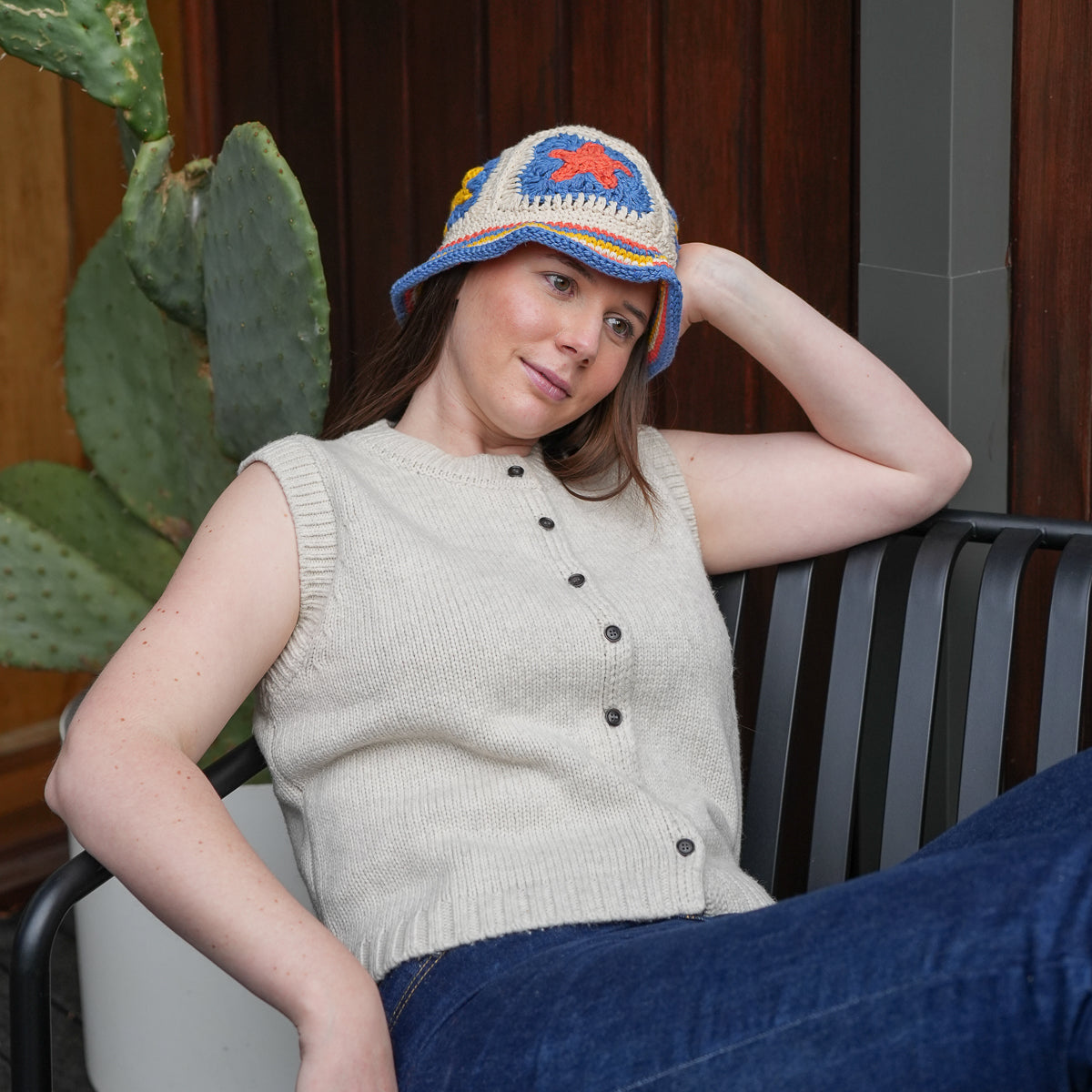 A woman sitting on a black metal patio chair, wearing a cream ribbed knit sleeveless vest with dark buttons, blue jeans, and a colorful crocheted bucket hat. A green cactus is visible in the background against a dark wood panel wall.