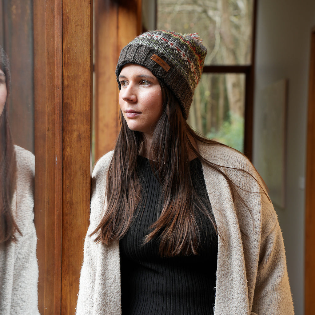 A woman wearing a multicolor knit beanie with a brown leather patch and a beige cardigan, standing in a warmly lit indoor setting with a wooden door and window in the background.