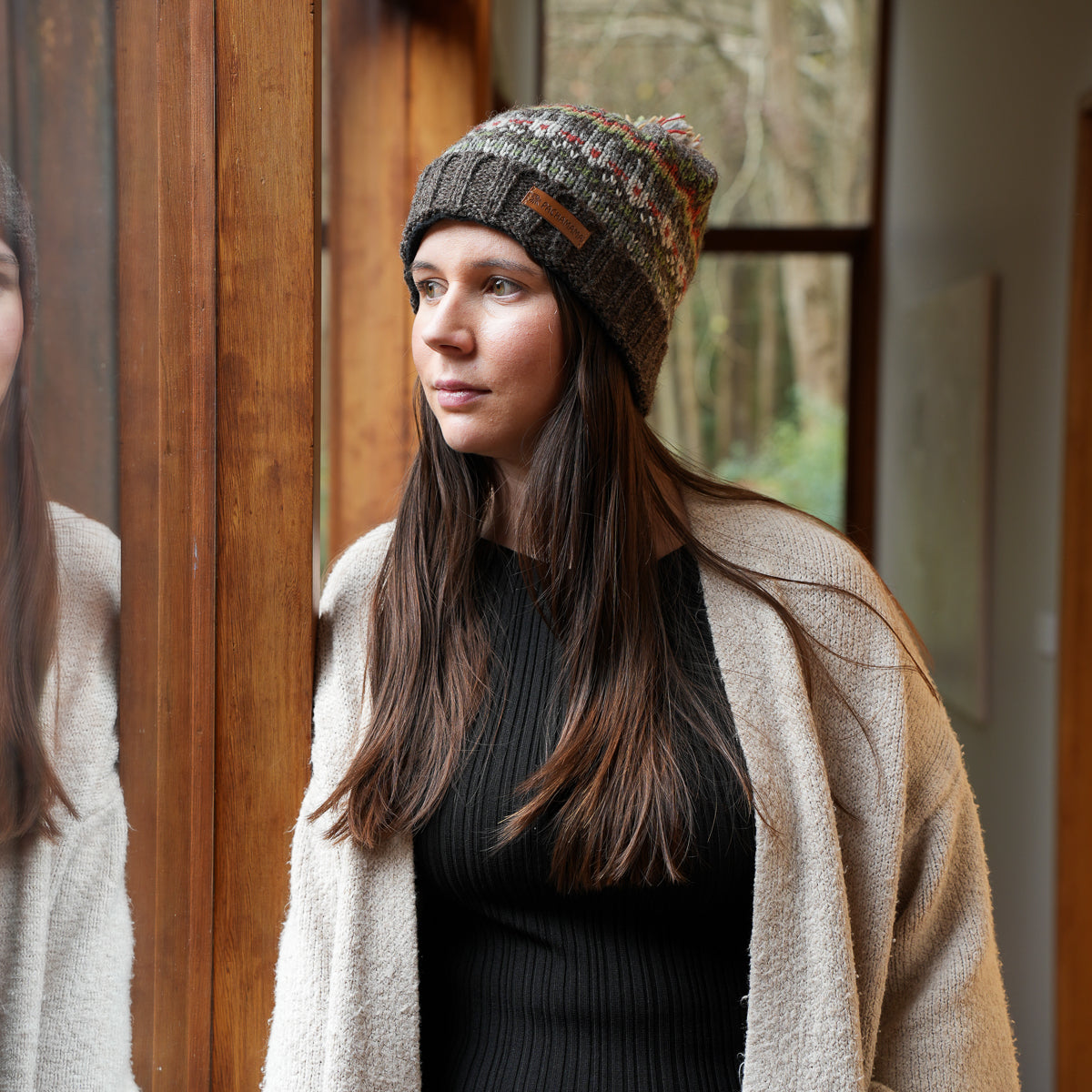 A woman wearing a multicolor knit beanie with a brown leather patch and a beige cardigan, standing in a warmly lit indoor setting with a wooden door and window in the background.