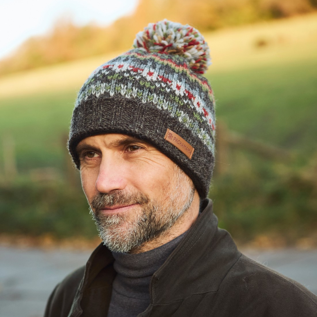 A man wearing a knit pom-pom beanie with a multicolor Fair Isle pattern and a brown leather brand tab, photographed outdoors against a blurred green hillside background.