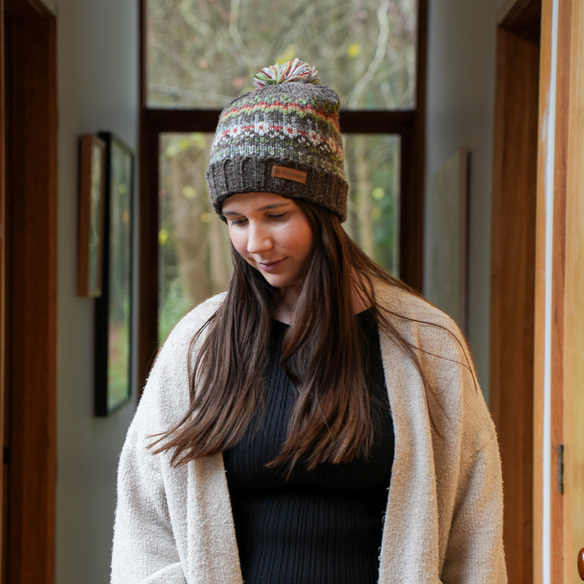 A woman wearing a multicolor knit pom-pom beanie hat and a cream cardigan, standing in a warmly lit indoor hallway.