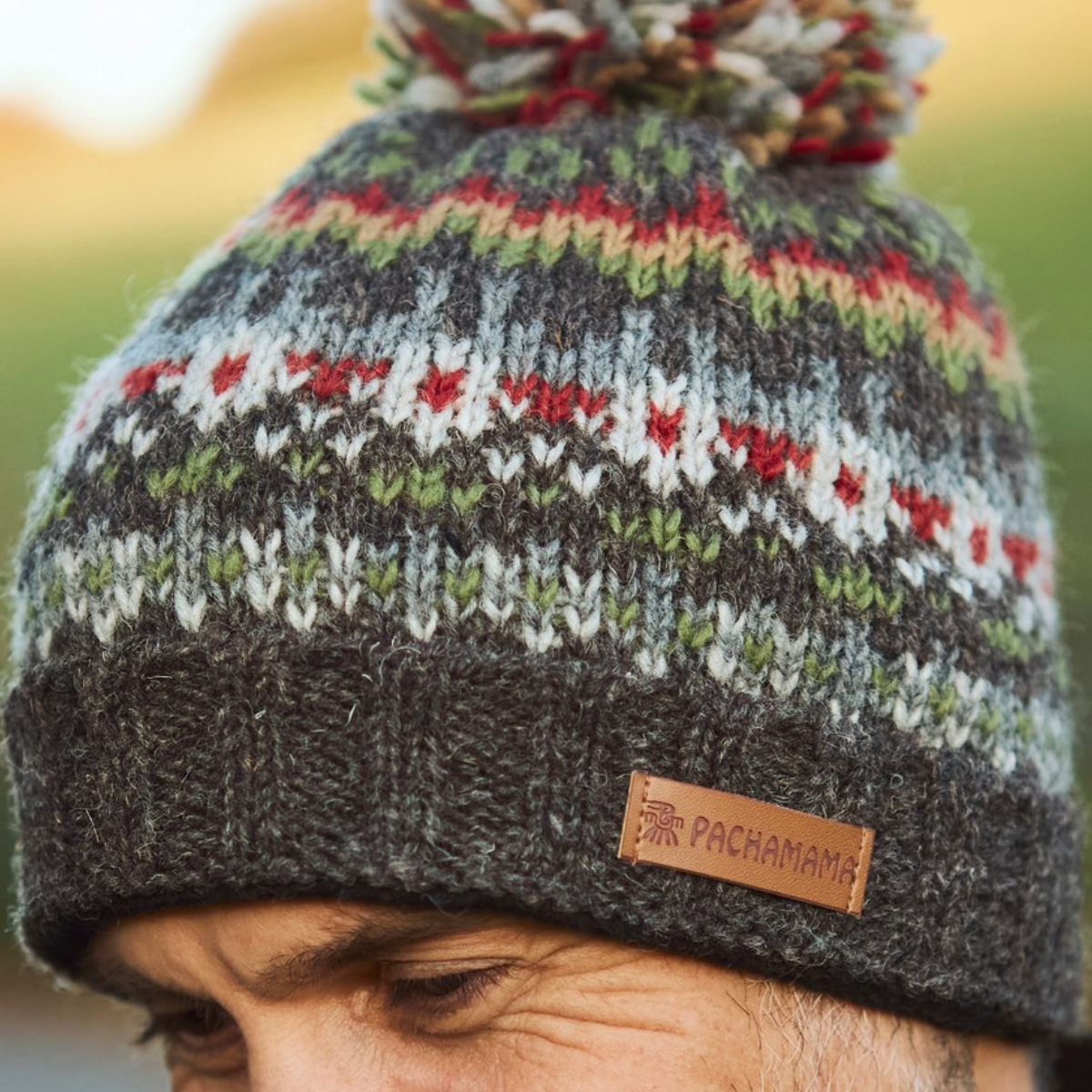 A close-up of a person wearing a knitted pom-pom beanie with horizontal multicolor stripes in gray, white, red, and green, featuring a dark gray ribbed cuff with a brown leather PACHAMAMA logo patch.