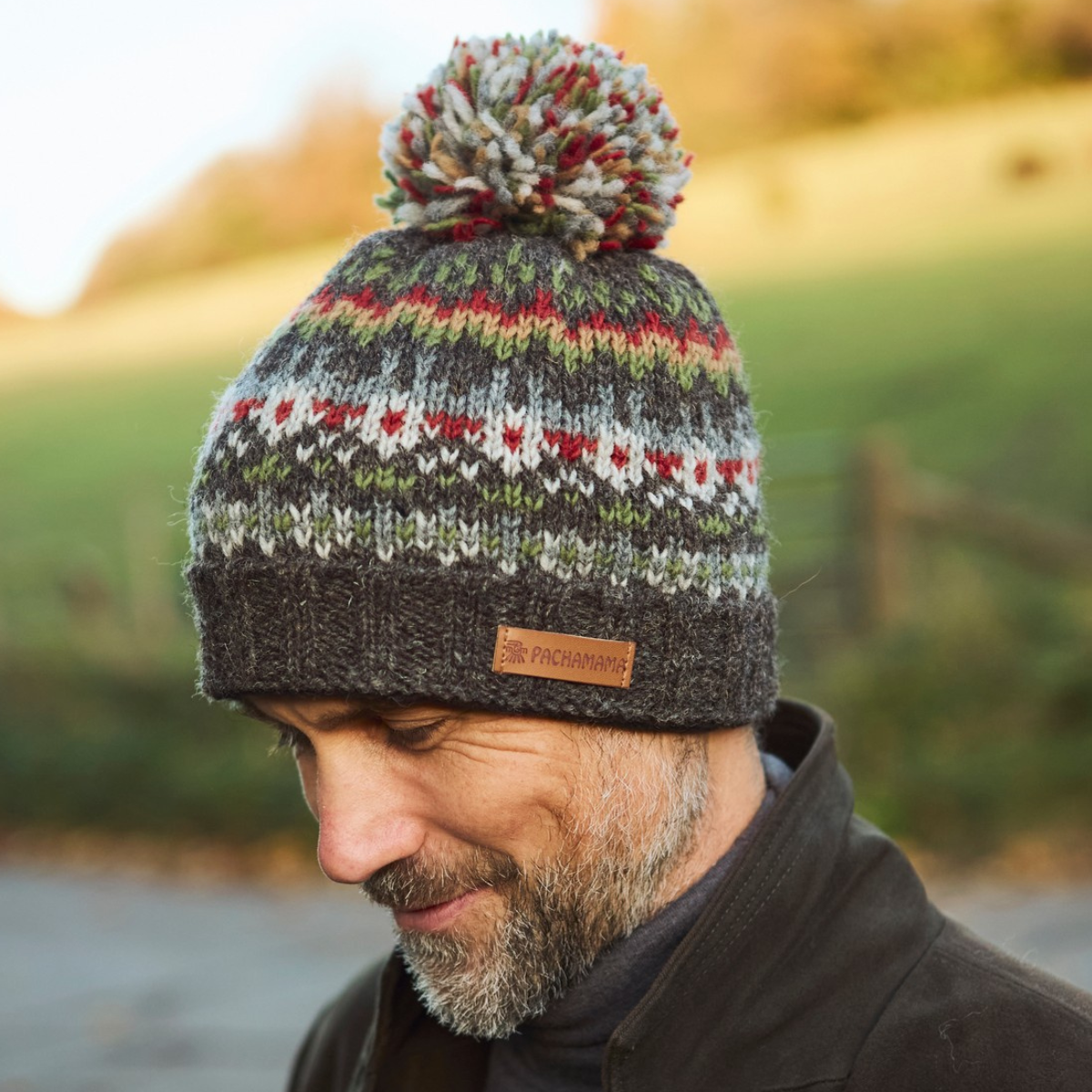 A man wearing a dark gray knit beanie with a multicolor Fair Isle pattern and a brown pom-pom. A small brown leather brand patch on the cuff reads 'FROGMARTIN'.