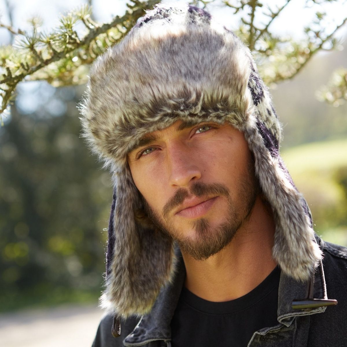 A man wearing a gray faux fur trapper hat with ear flaps and a dark denim jacket, photographed outdoors with blurred trees in the background.